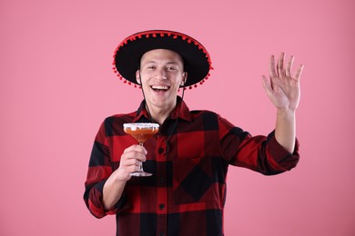 Photo of Smiling man in black Flamenco hat with cocktail waving hello on pink background
