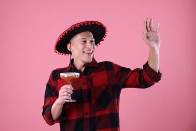 Photo of Smiling man in black Flamenco hat with cocktail waving hello on pink background