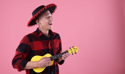 Photo of Smiling man in black Flamenco hat playing ukulele on pink background. Space for text