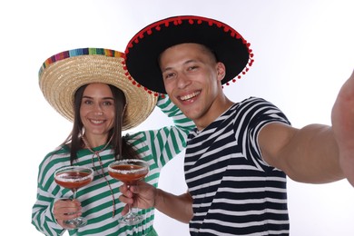 Photo of Happy couple in sombrero and Flamenco hats with cocktails taking selfie on white background