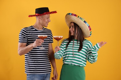 Photo of Happy couple in sombrero and Flamenco hats with cocktails on yellow background