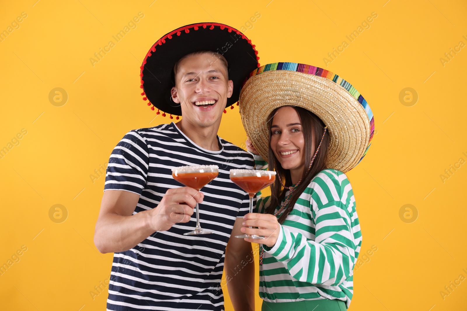 Photo of Happy couple in sombrero and Flamenco hats with cocktails on yellow background