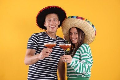 Photo of Happy couple in sombrero and Flamenco hats with cocktails on yellow background