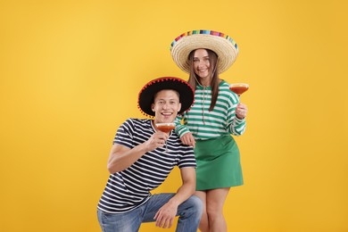 Photo of Happy couple in sombrero and Flamenco hats with cocktails on yellow background
