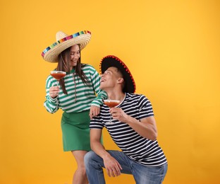 Photo of Happy couple in sombrero and Flamenco hats with cocktails on yellow background
