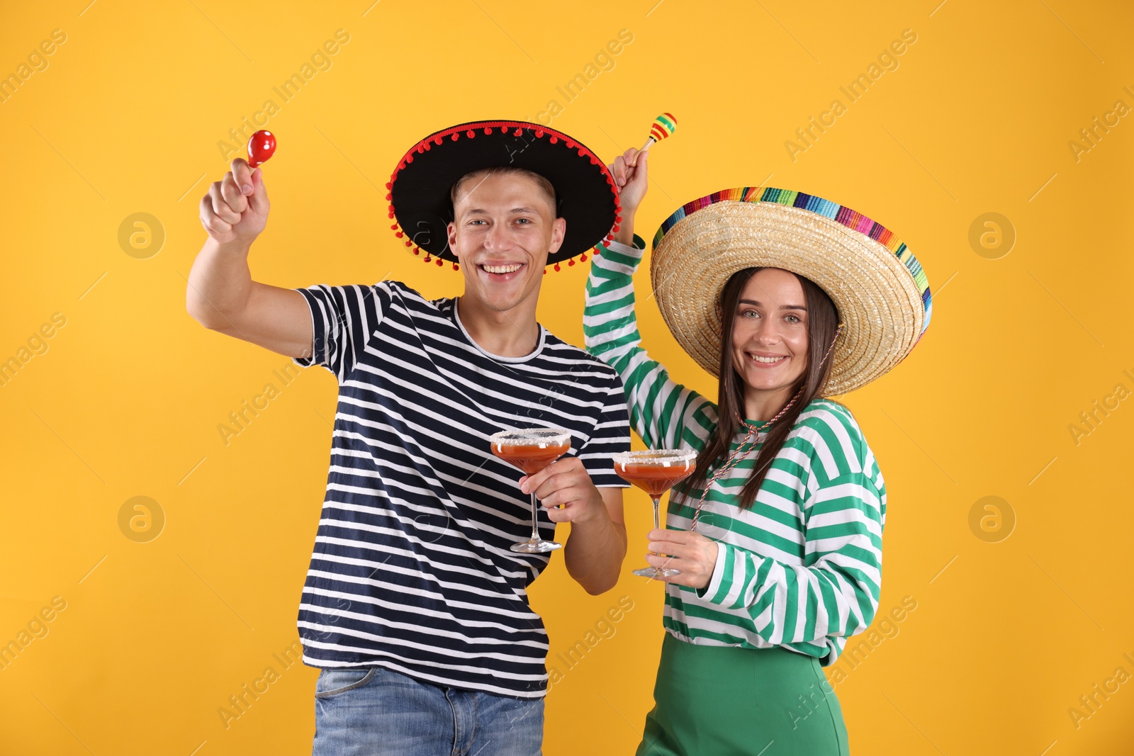 Photo of Happy couple in sombrero and Flamenco hats. Man and woman with cocktails and maracas on yellow background