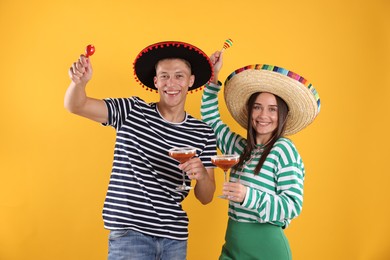 Photo of Happy couple in sombrero and Flamenco hats. Man and woman with cocktails and maracas on yellow background