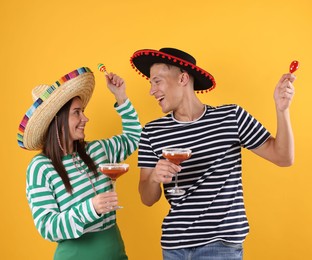 Photo of Happy couple in sombrero and Flamenco hats. Man and woman with cocktails and maracas on yellow background