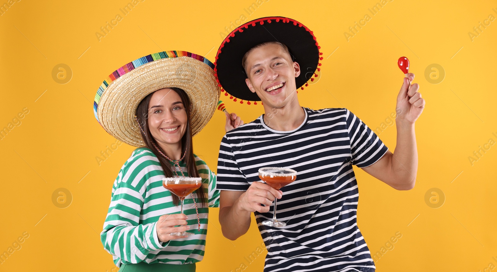 Photo of Happy couple in sombrero and Flamenco hats. Man and woman with cocktails and maracas on yellow background