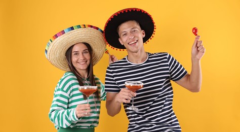 Photo of Happy couple in sombrero and Flamenco hats. Man and woman with cocktails and maracas on yellow background