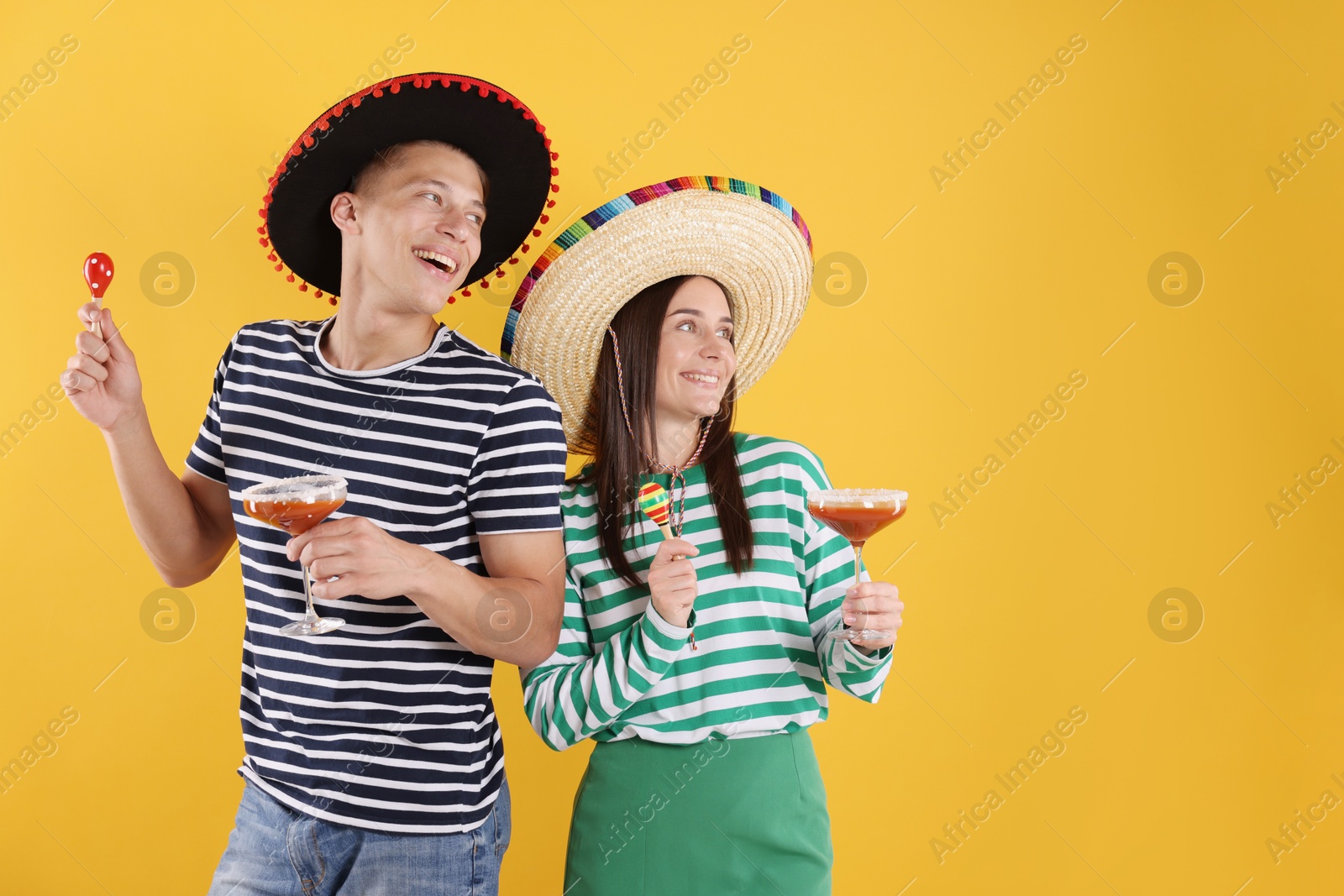 Photo of Happy couple in sombrero and Flamenco hats. Man and woman with cocktails and maracas on yellow background. Space for text