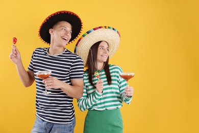 Photo of Happy couple in sombrero and Flamenco hats. Man and woman with cocktails and maracas on yellow background. Space for text