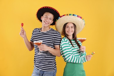 Photo of Happy couple in sombrero and Flamenco hats. Man and woman with cocktails and maracas on yellow background