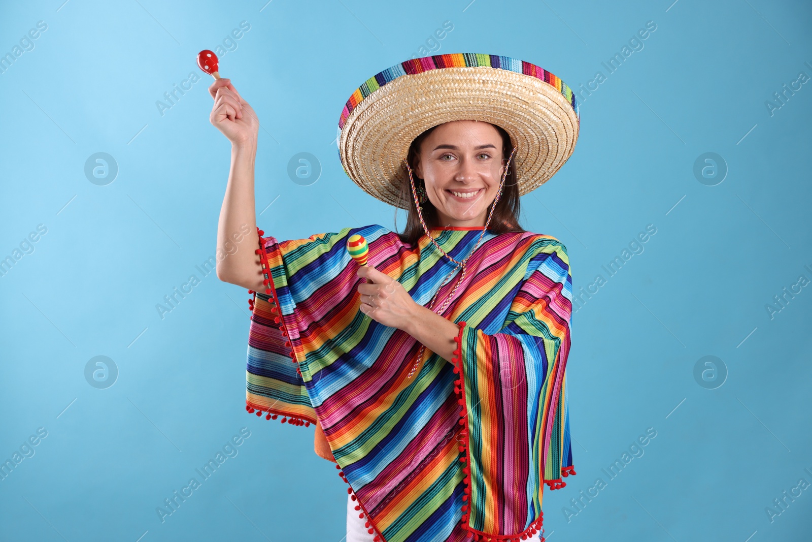 Photo of Happy woman in sombrero hat and colorful poncho playing maracas on light blue background