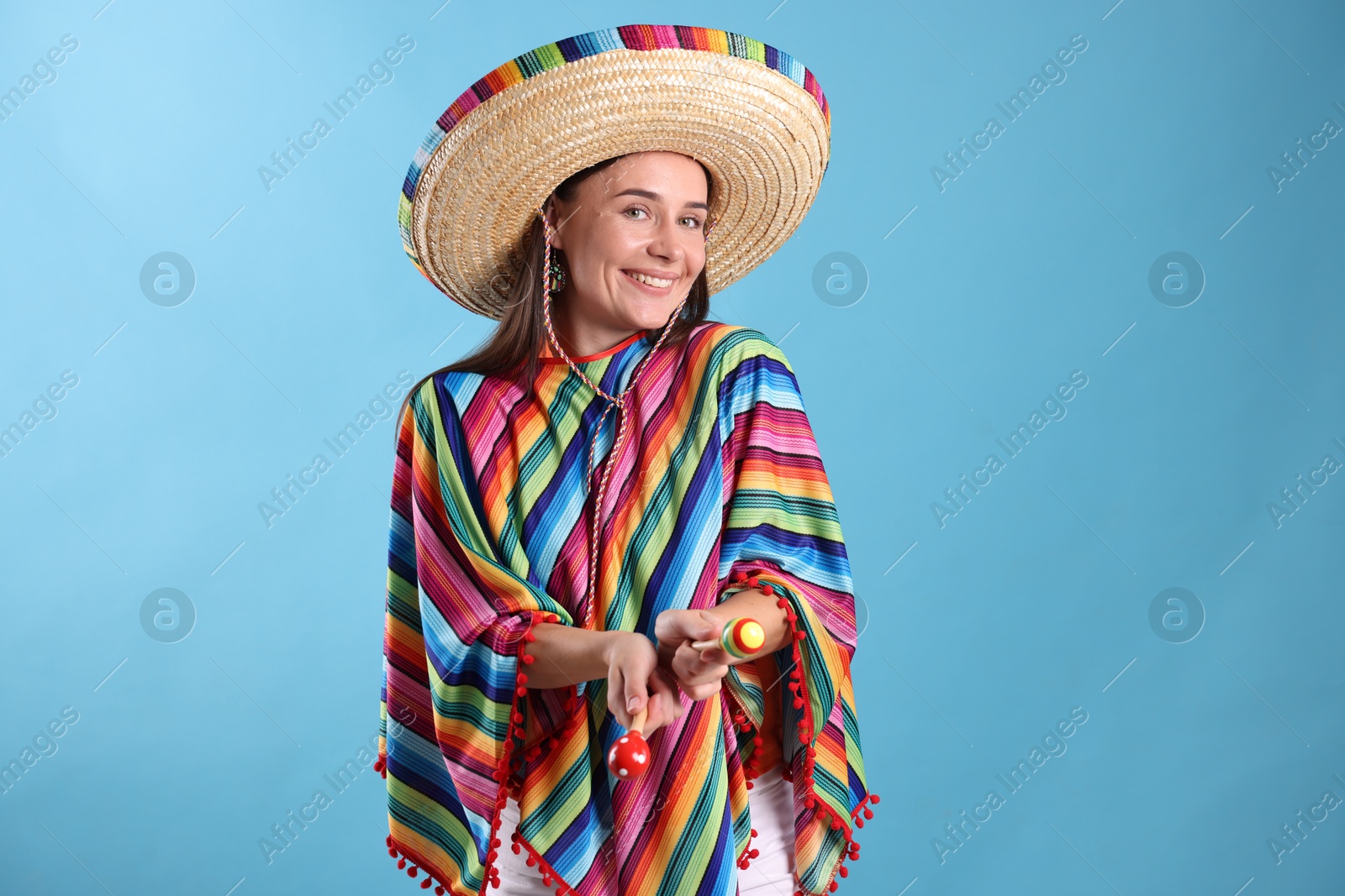 Happy woman in sombrero hat and colorful poncho playing maracas on light blue background Photo of Happy woman in sombrero hat and colorful poncho playing maracas on light blue background