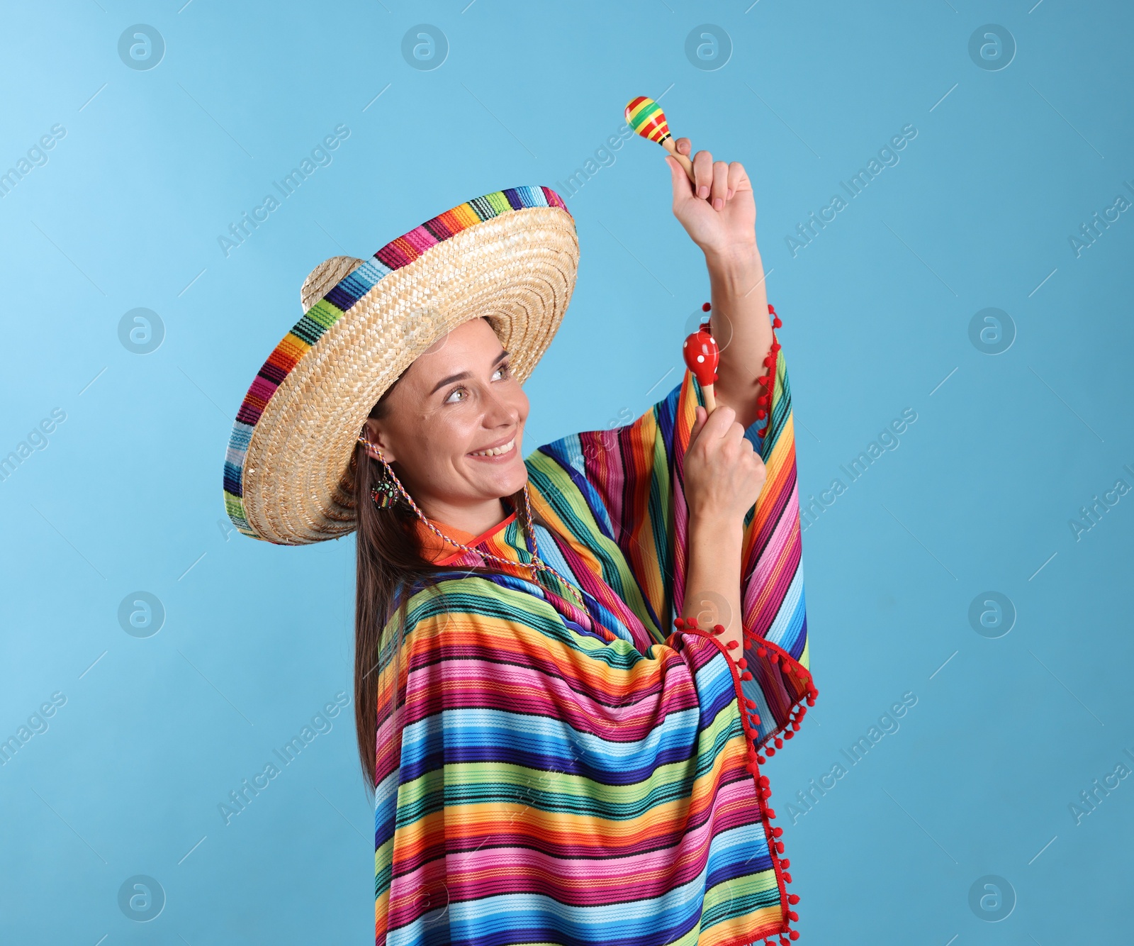 Photo of Happy woman in sombrero hat and colorful poncho playing maracas on light blue background