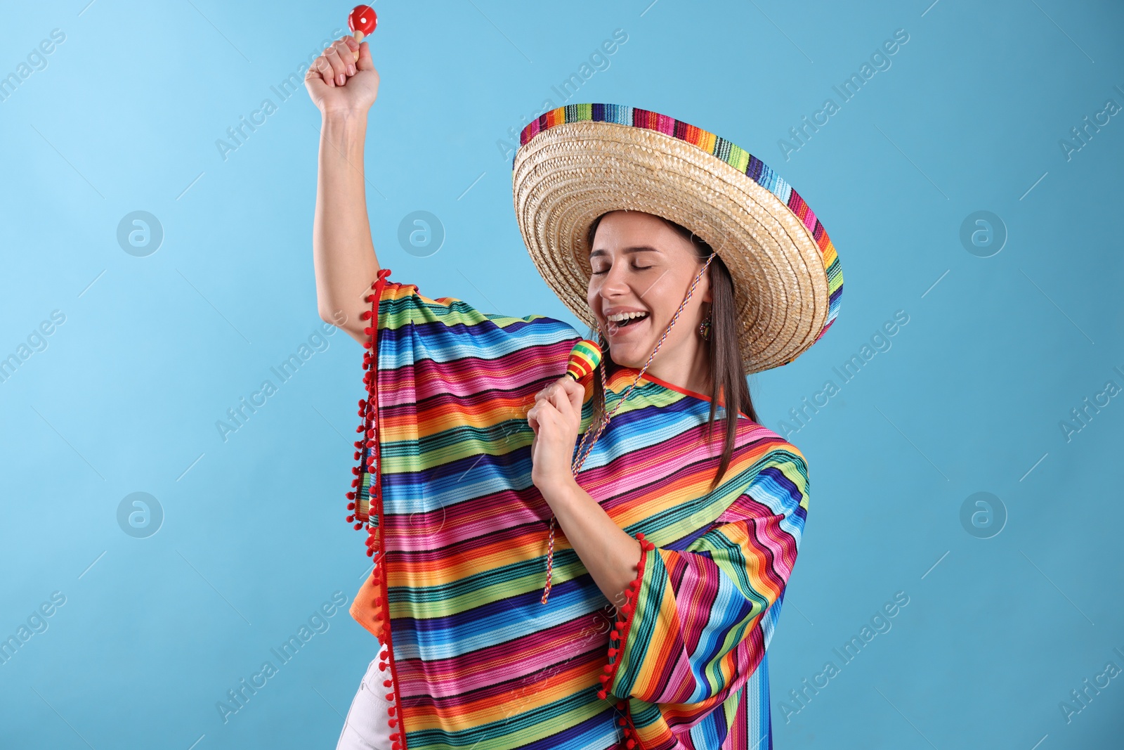 Photo of Happy woman in sombrero hat and colorful poncho playing maracas on light blue background