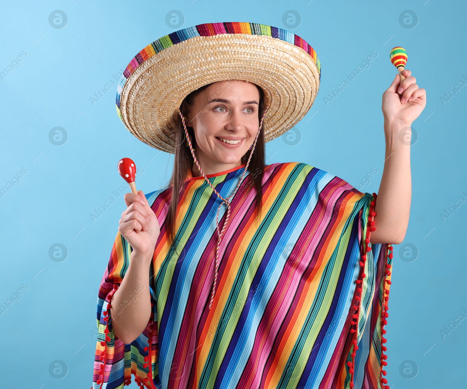 Happy woman in sombrero hat and colorful poncho playing maracas on light blue background Photo of Happy woman in sombrero hat and colorful poncho playing maracas on light blue background