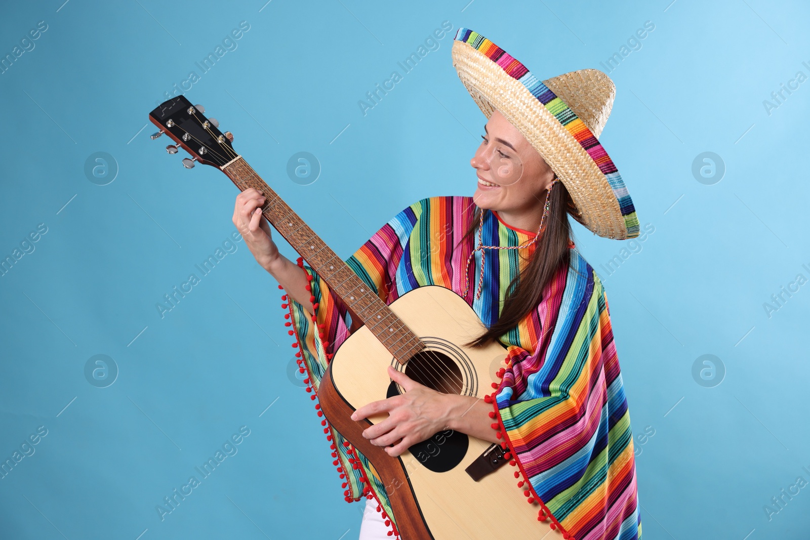 Smiling woman in sombrero hat and colorful poncho playing guitar on light blue background Photo of Smiling woman in sombrero hat and colorful poncho playing guitar on light blue background