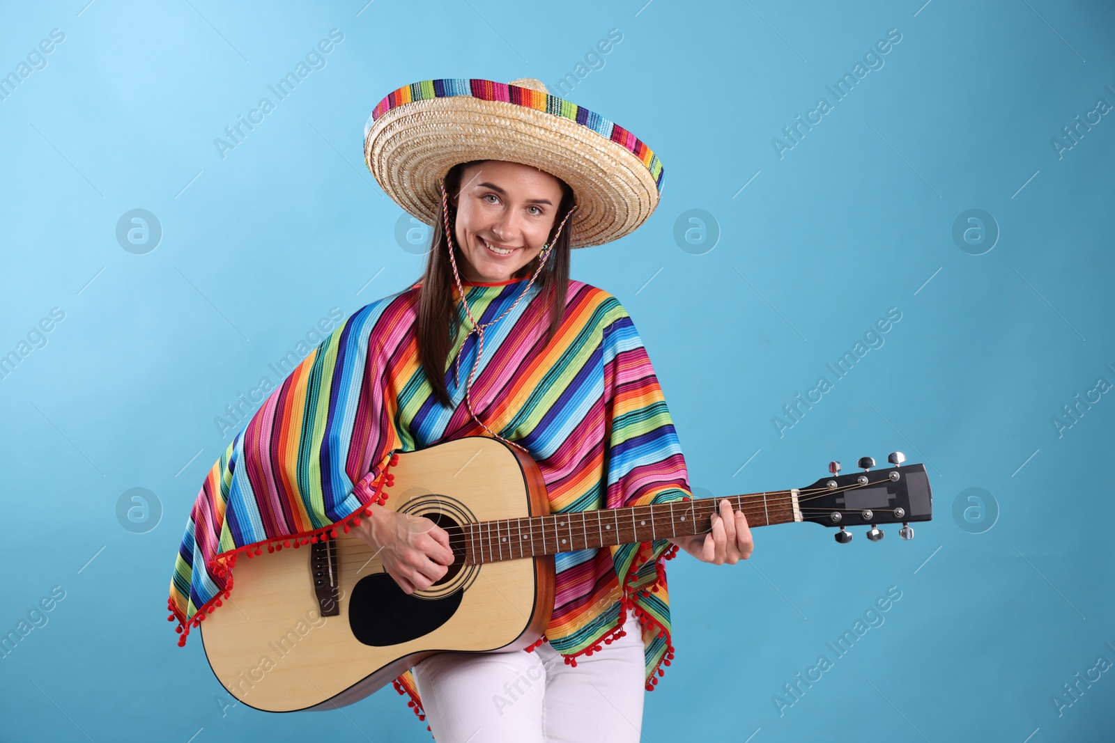 Photo of Smiling woman in sombrero hat and colorful poncho playing guitar on light blue background