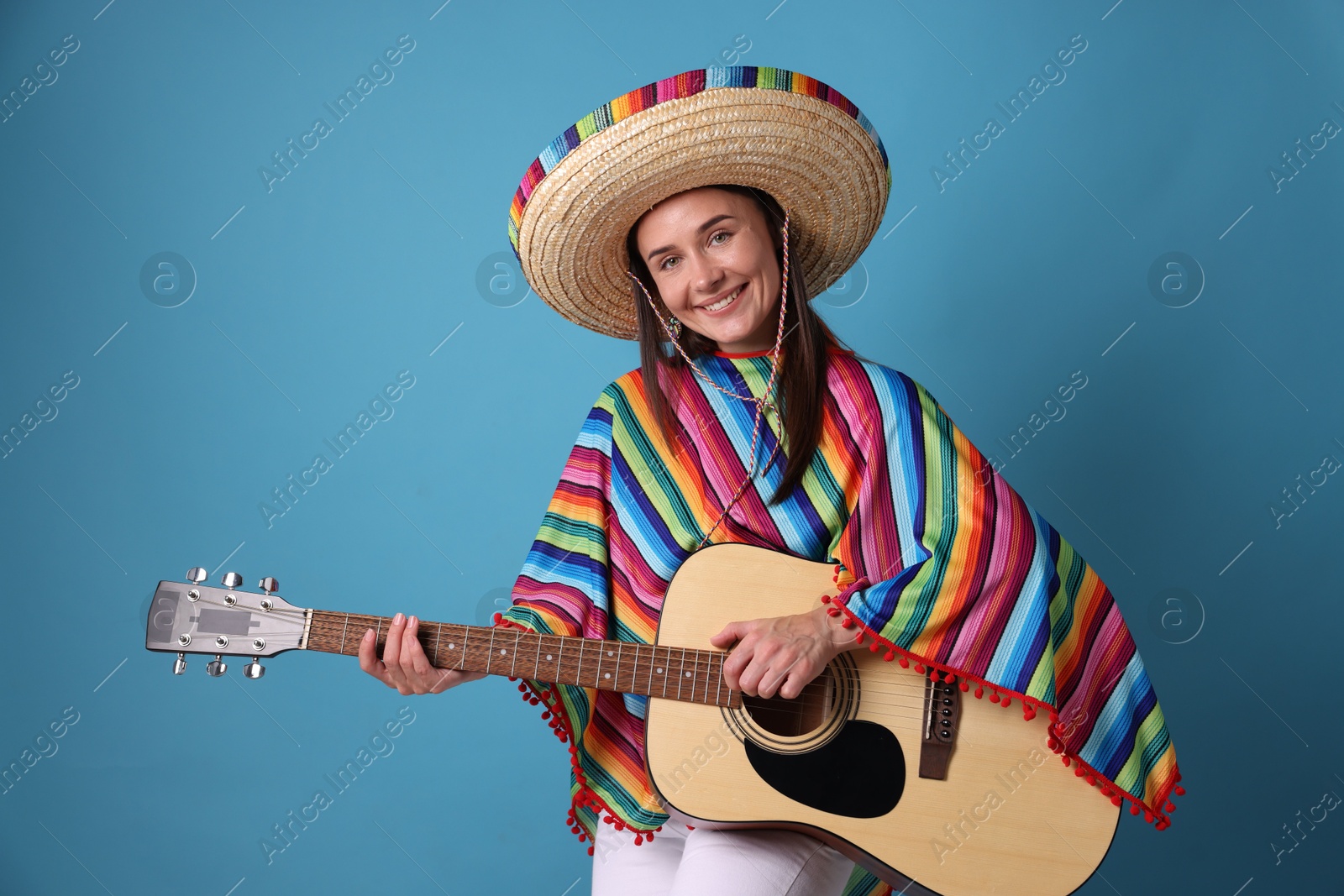 Photo of Smiling woman in sombrero hat and colorful poncho playing guitar on light blue background