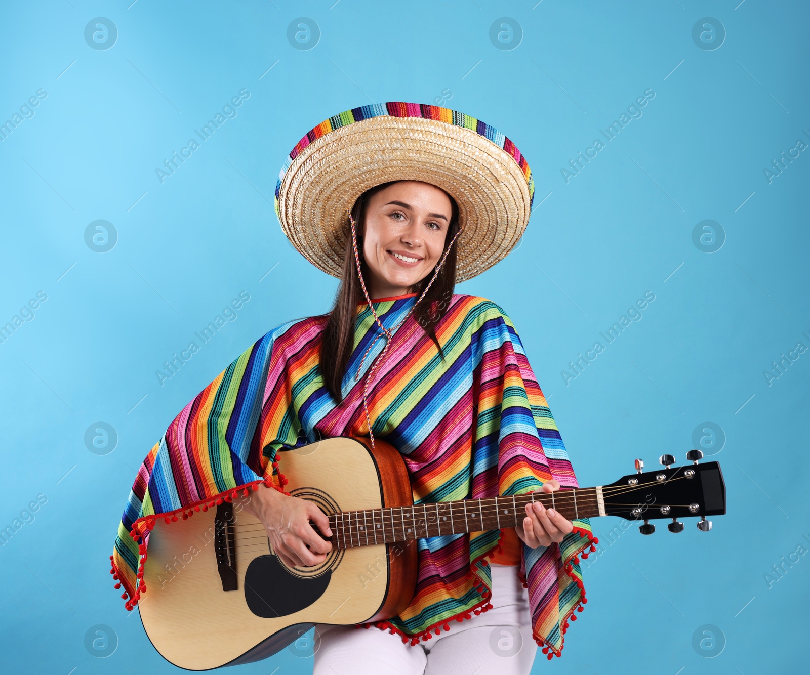 Smiling woman in sombrero hat and colorful poncho playing guitar on light blue background Photo of Smiling woman in sombrero hat and colorful poncho playing guitar on light blue background