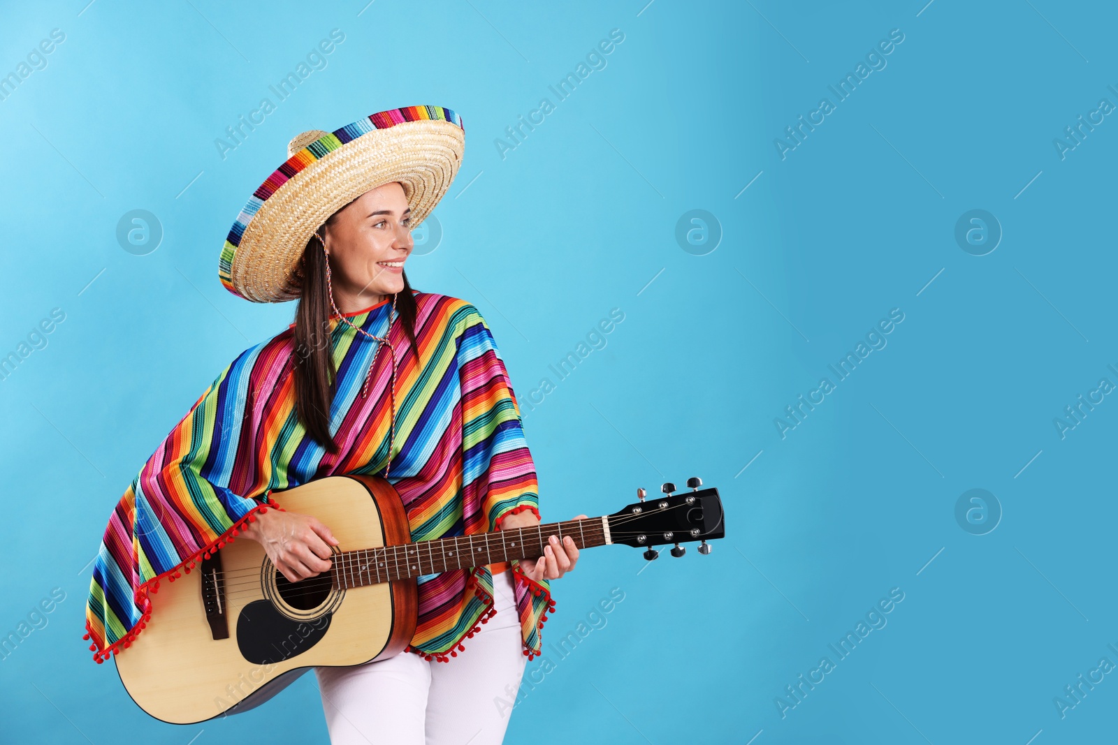 Photo of Smiling woman in sombrero hat and colorful poncho playing guitar on light blue background. Space for text