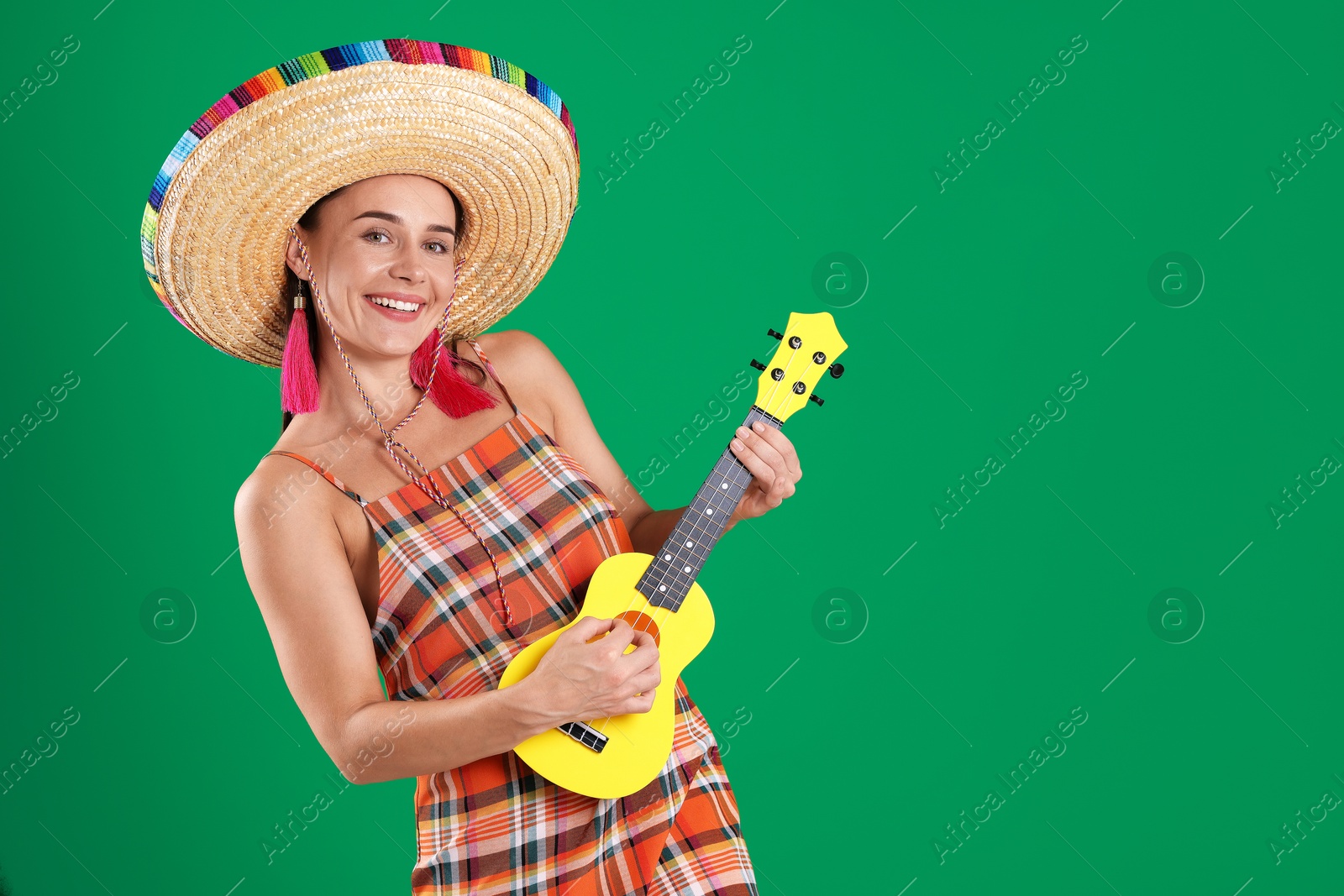 Photo of Smiling woman in sombrero hat playing ukulele on green background. Space for text