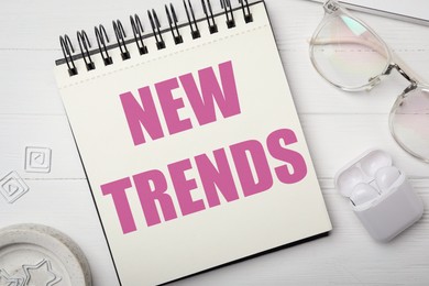 Image of New Trends inscription in notebook, glasses and earphones on white wooden table, top view