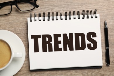 Image of Trends inscription in notebook, coffee, glasses and pen on wooden table, top view
