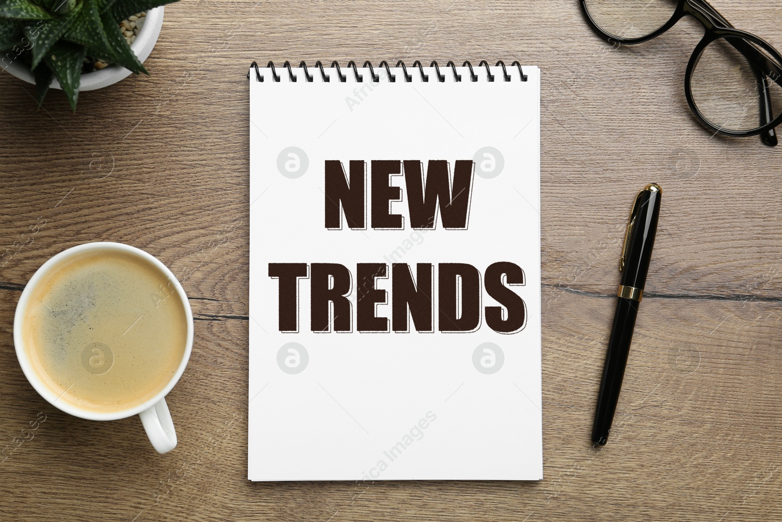 New Trends inscription in notebook, coffee, glasses and pen on wooden table, top view Image of New Trends inscription in notebook, coffee, glasses and pen on wooden table, top view