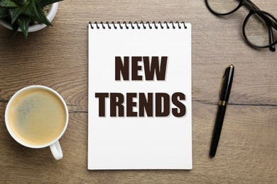 Image of New Trends inscription in notebook, coffee, glasses and pen on wooden table, top view