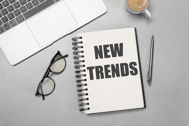 Image of New Trends inscription in notebook, laptop, coffee, glasses and pen on grey table, top view
