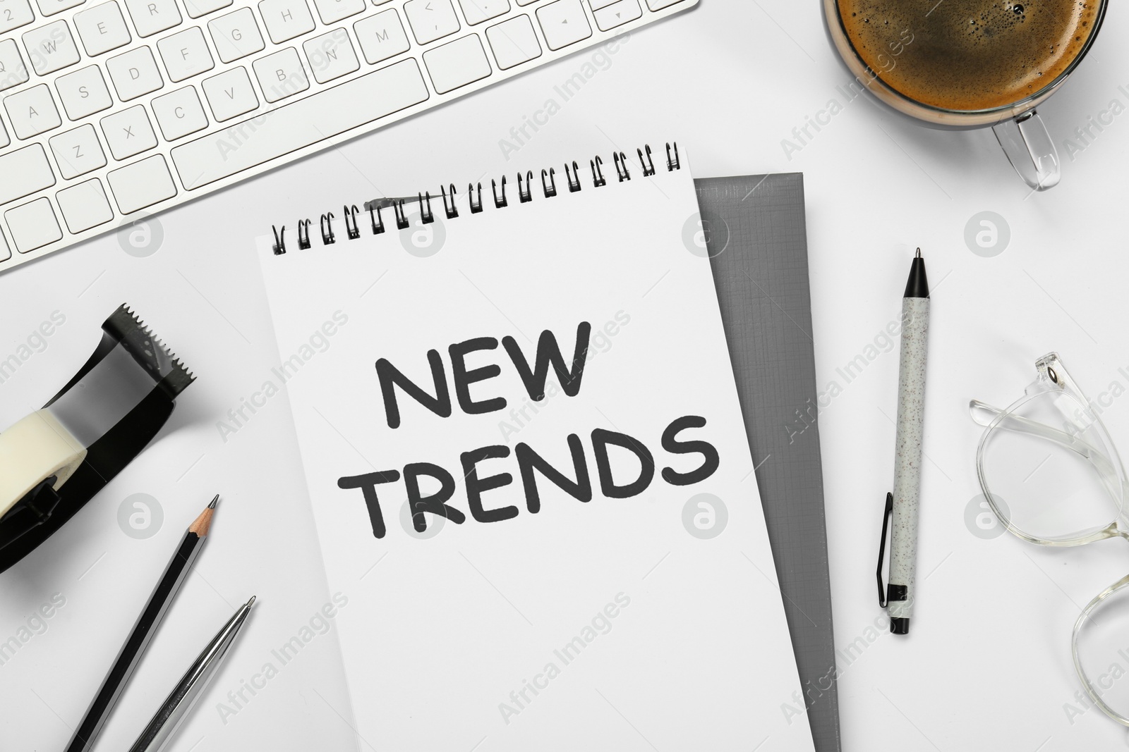 New Trends inscription in notebook, coffee, computer keyboard and stationery on white table, top view Image of New Trends inscription in notebook, coffee, computer keyboard and stationery on white table, top view