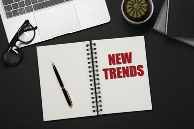 Image of New Trends inscription in notebook, laptop, glasses and pen on black table, top view