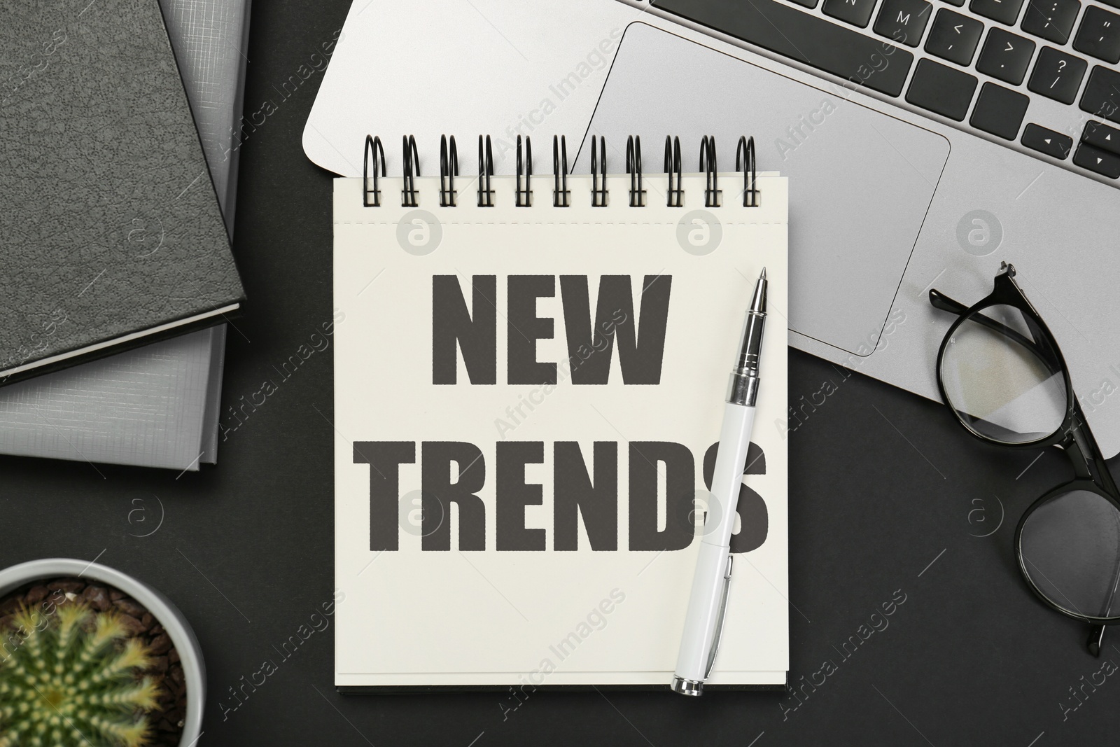 Image of New Trends inscription in notebook, laptop, glasses and pen on black table, top view