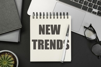 Image of New Trends inscription in notebook, laptop, glasses and pen on black table, top view