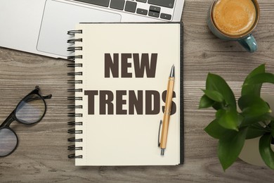 Image of New Trends inscription in notebook, coffee, glasses, laptop and pen on wooden table, top view