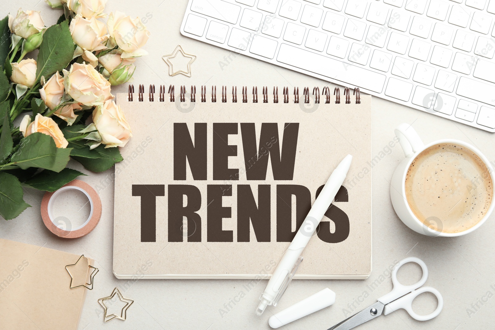 New Trends inscription in notebook, computer keyboard, flowers, coffee and stationery on beige table, top view Image of New Trends inscription in notebook, computer keyboard, flowers, coffee and stationery on beige table, top view