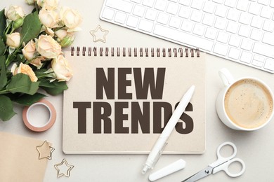 Image of New Trends inscription in notebook, computer keyboard, flowers, coffee and stationery on beige table, top view