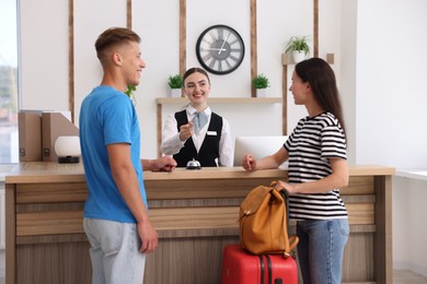 Photo of Smiling receptionist in uniform giving hotel key card to couple of tourists at reception desk indoors