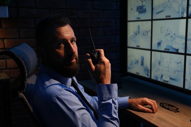Photo of Security guard monitoring CCTV cameras and using walkie talkie in office at night