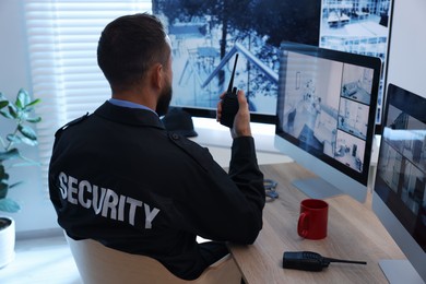 Photo of Security guard monitoring CCTV cameras and using walkie talkie in office, back view