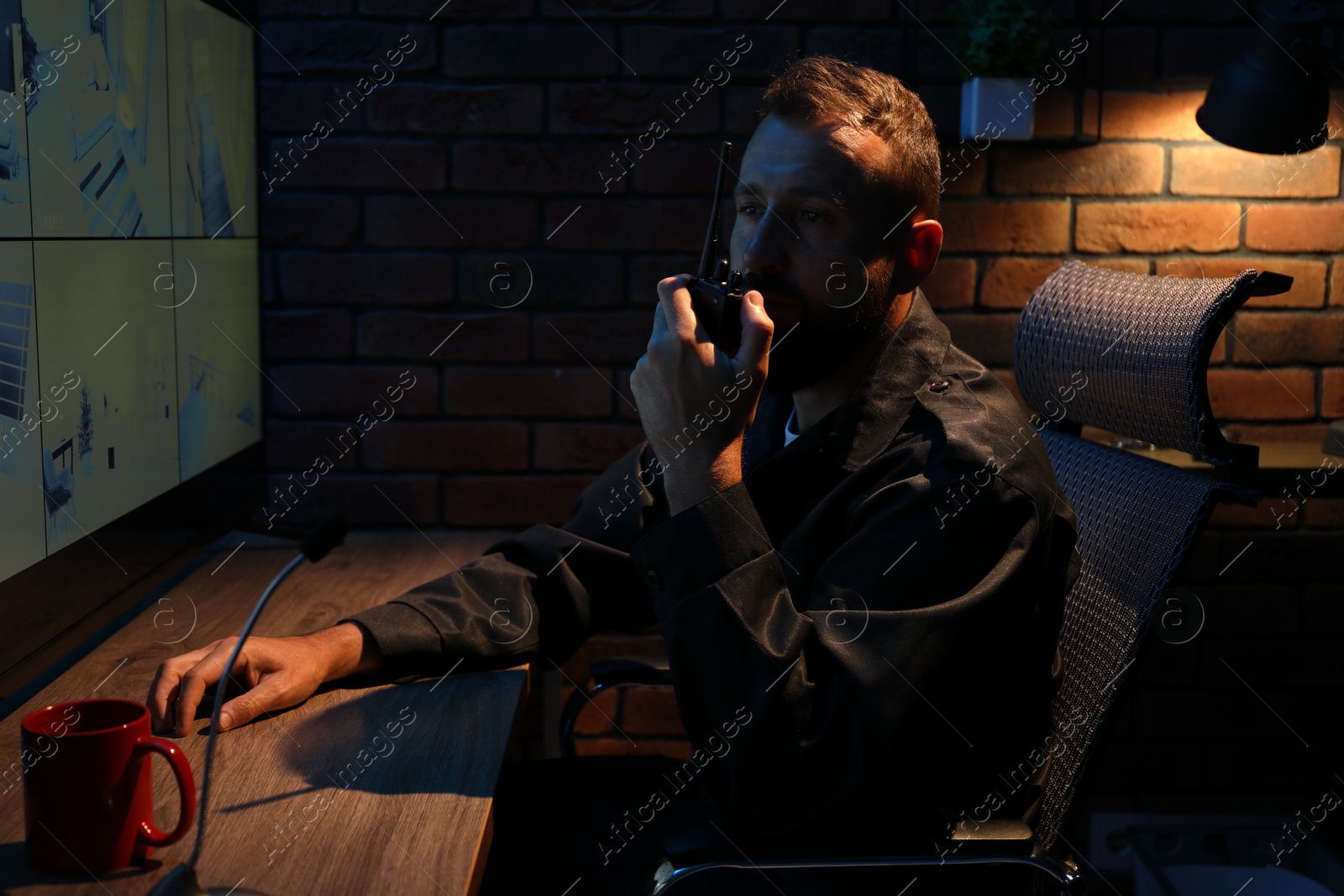 Photo of Security guard monitoring CCTV cameras and using walkie talkie in office at night