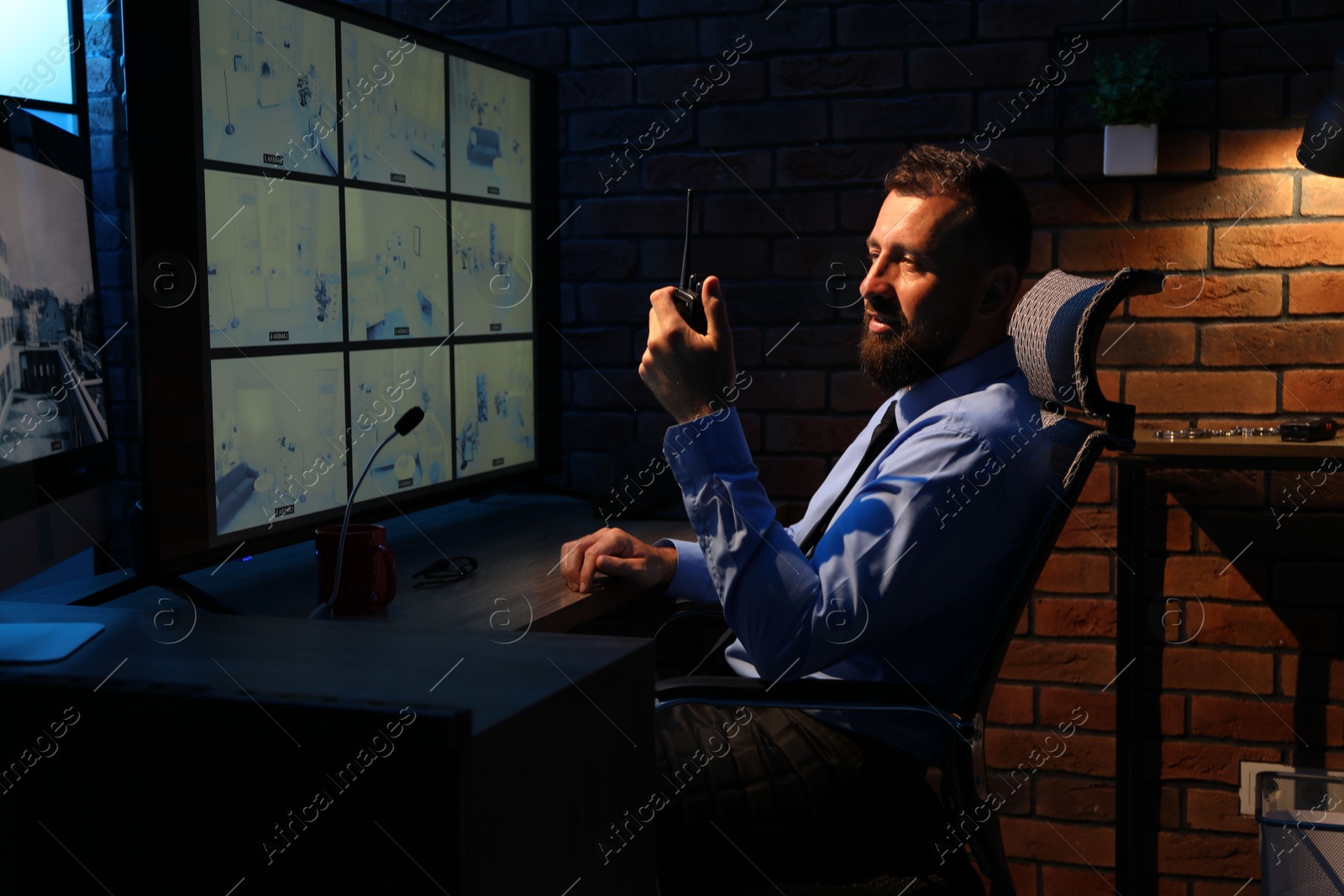 Photo of Security guard monitoring CCTV cameras and using walkie talkie in office at night