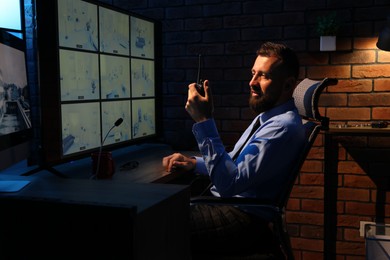 Photo of Security guard monitoring CCTV cameras and using walkie talkie in office at night