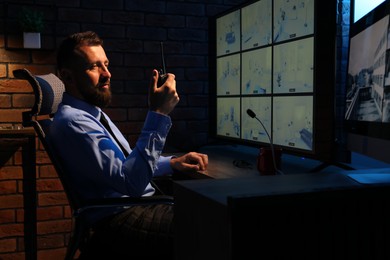 Photo of Security guard monitoring CCTV cameras and using walkie talkie in office at night