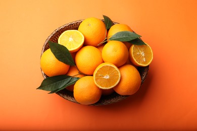 Photo of Fresh ripe oranges and green leaves in wicker basket on orange background, top view