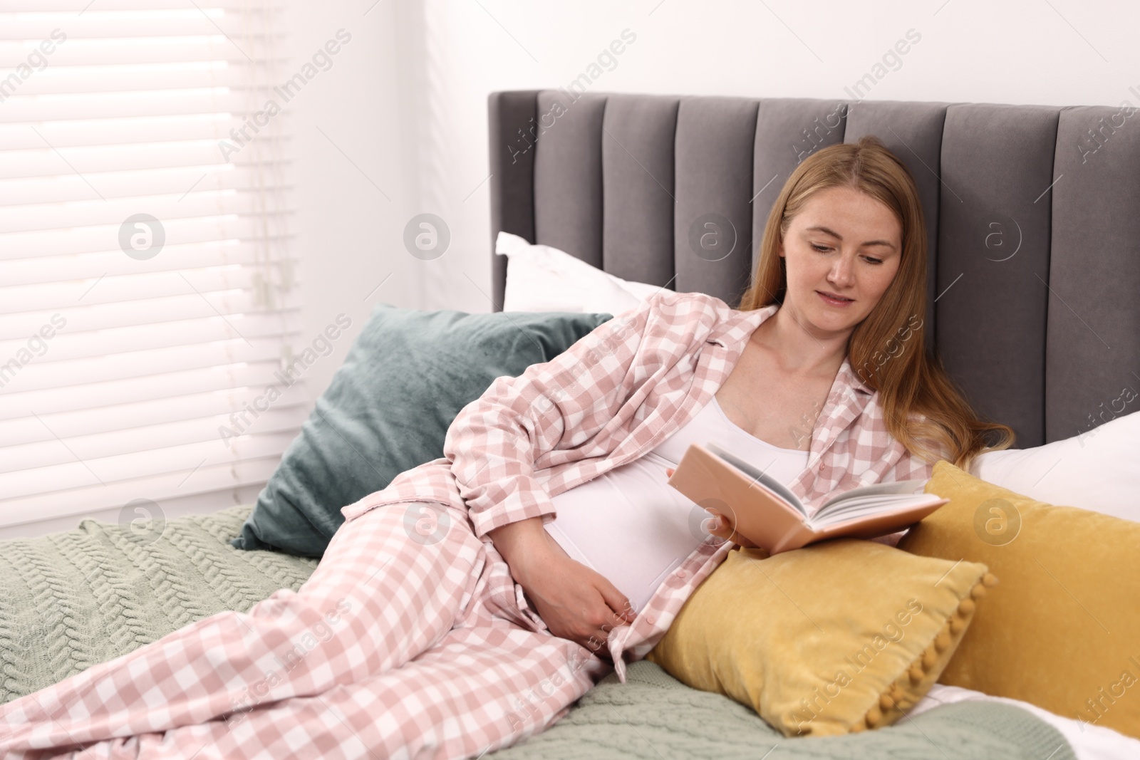Photo of Beautiful pregnant woman reading book on bed at home. Space for text