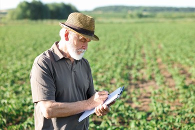 Photo of Senior farmer with clipboard in field of unripe soy outdoors, space for text
